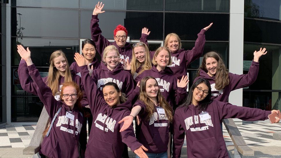 A dozen young women dressed in matching purple hoodies pose, arms in the air, for the camera