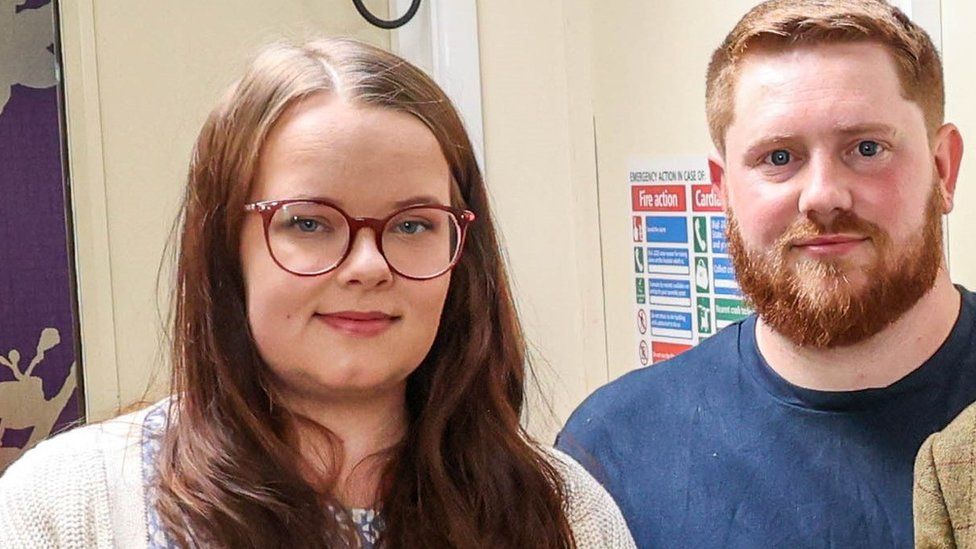 Tamar Vellacott and Cameron Pratt looking at the camera. Tamar wears glasses and has long hair, Cameron has ginger hair and a beard.