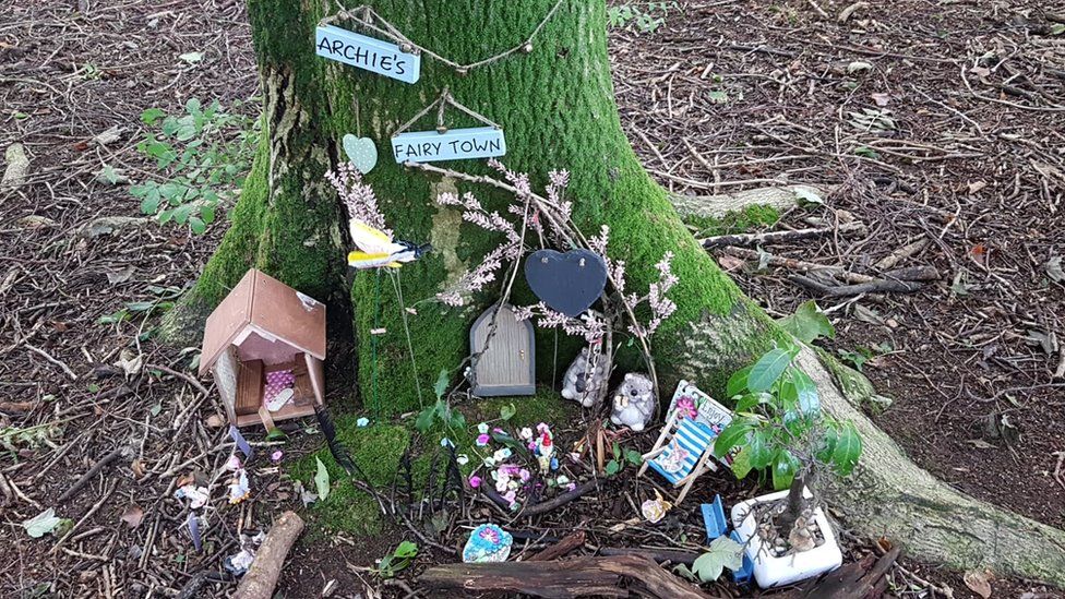 A miniature door, house and decorations at the base of a tree with a sign for "Archie's fairy town"