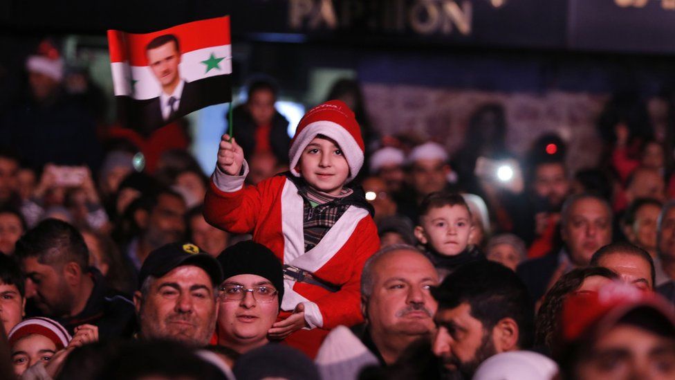 A child waves a flag of Syrian President while wearing a santa costume