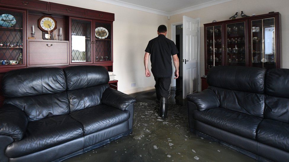 Man in a flooded living room wearing wellies and walking towards the exit door