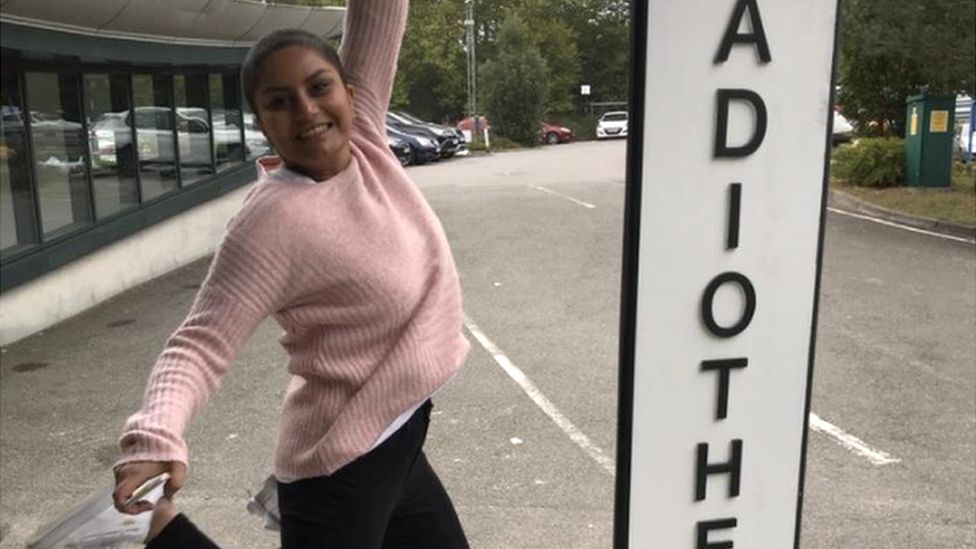 Sania is wearing a pink sweater and black trousers. She is jumping up smiling at the camera, with the sign to the hospital next to her. The background is of a car park, with glass buidlings on the left and cars directly behind, with trees and leaves overhanging.
