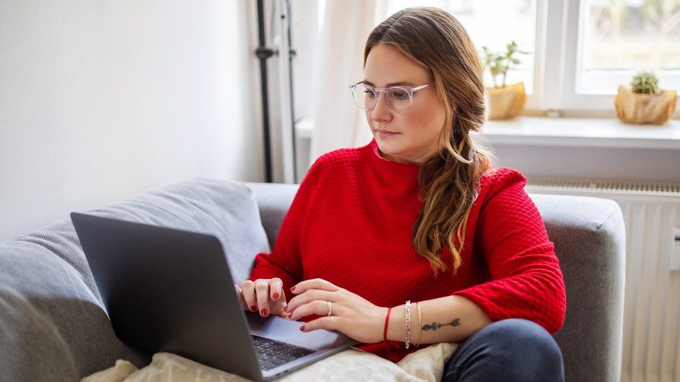 A woman in a red jumper sits at her laptop on a sofa