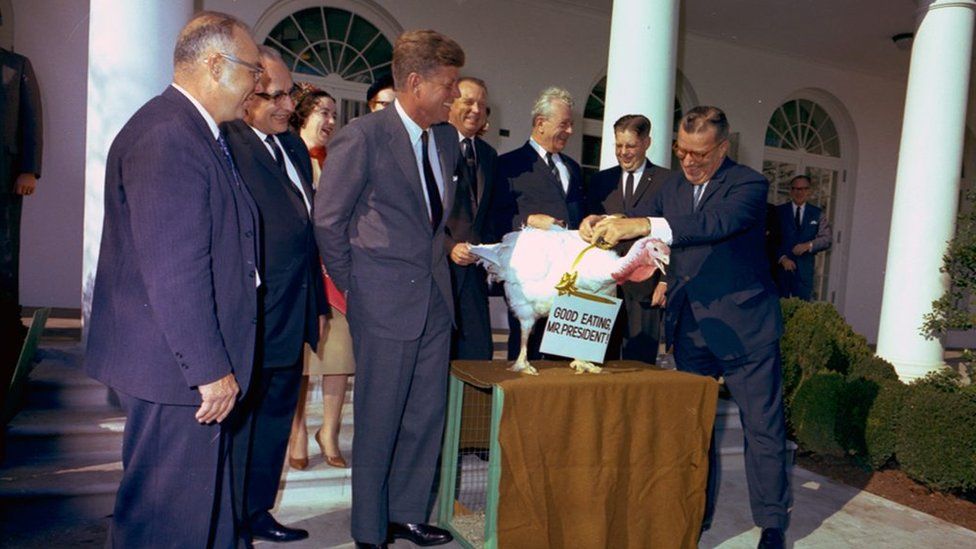 JFK stands with administration officials while a turkey is presented saying 'Good Eating Mr President' around its neck