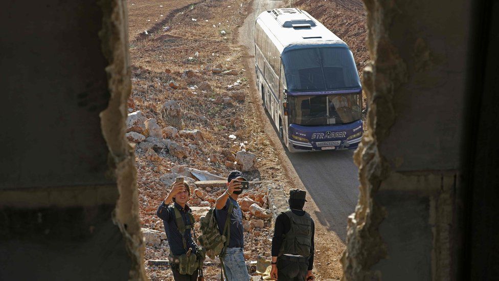 Members of Hayat Tahrir al-Sham group, led by Syria's former Al-Qaeda affiliate, stand as a convoy of busses gets ready to enter the towns of Fouaa and Kafraya to evacuate their residents, 18 July 2018