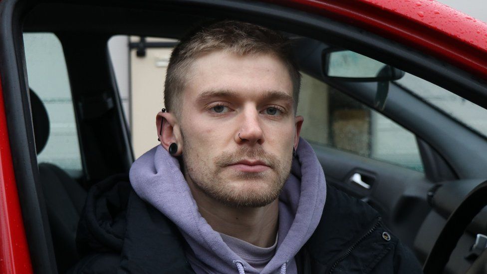 A young man sits in the driver's seat of a red saloon car, with the door open and him facing outwards. He's got an impassive look on his face and short, blonde hair and stubble. He has a black flesh tunnel piercing in his right earlobe, and an industrial bar piercing - also black - across the top of his ear. He wears a black jacket over a lilac hoodie.