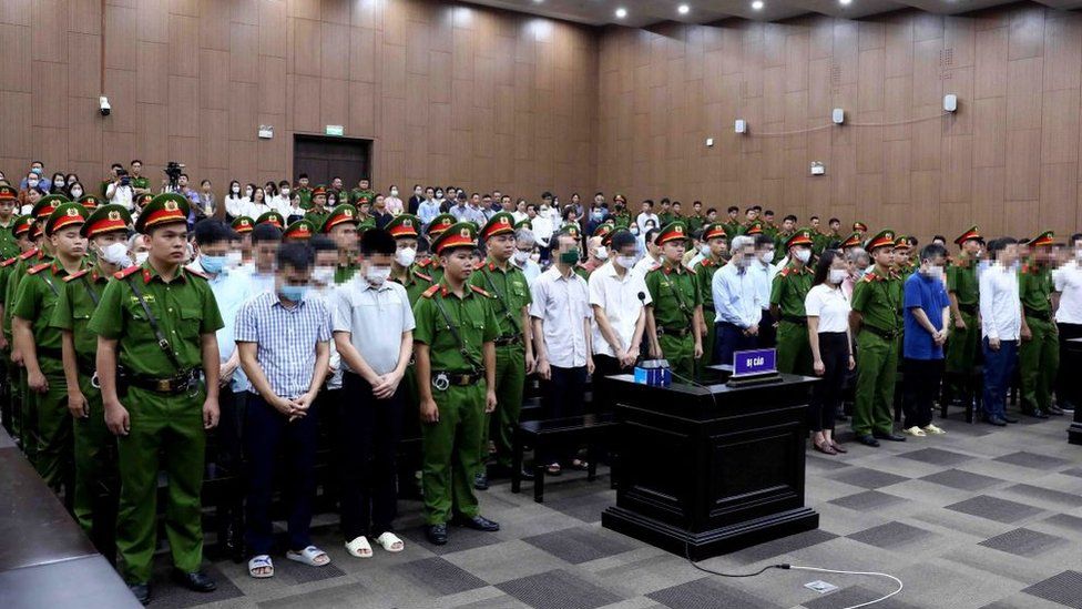 На этом снимке, сделанном и опубликованном Вьетнамским информационным агентством 28 июля 2023 года, изображены обвиняемые (в гражданских рубашках), стоящие перед вынесением приговора в зале суда Ханоя по делу о репатриации.