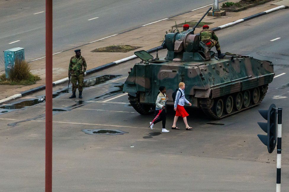 Young women walk past an armoured personnel carrier.