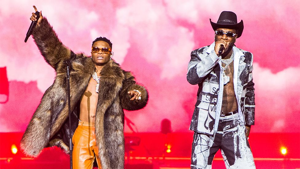 Wizkid (left) and Burna Boy (right) perform together. Wizkid is wearing a brown fur coat, with orange leather trousers and heavy chains around his neck. His right arm is pointing to the sky with his hand holding a microphone. He is wearing sunglasses with his left arm pointing to the crowd. Burna Boy is wearing a black and white top and bottom combination, with a hat and sunglasses, as his right arm is holding a microphone which he is singing into. The background is smoky and coloured red, with floor spotlights shining up.