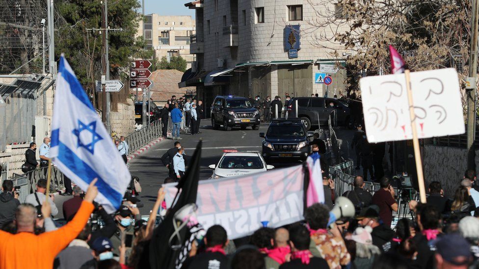 Protesters hold up placards and an Israeli flag as a convoy transporting Israeli Prime Minister Benjamin Netanyahu arrives at the Jerusalem District Court for a hearing on 8 February 2021