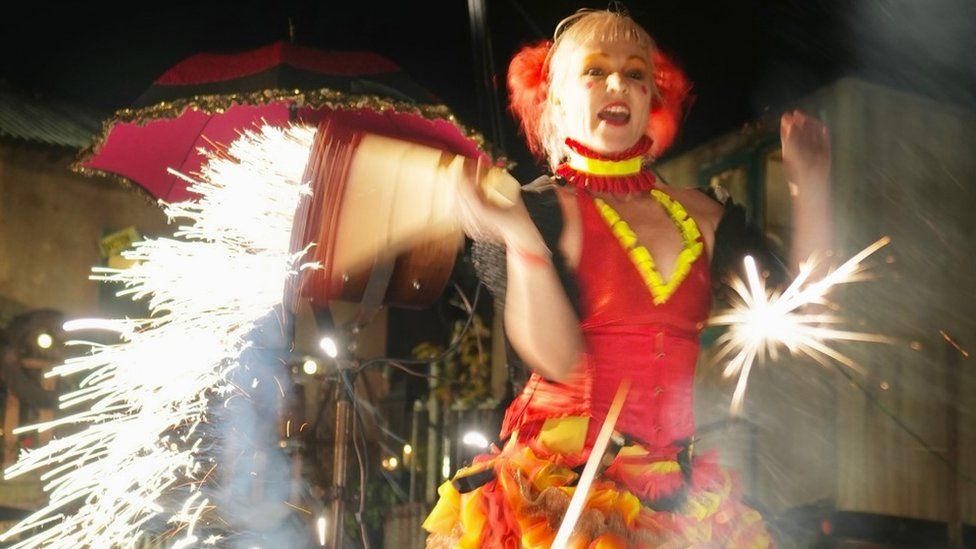A circus performer in red, yellow and gold with light blonde hair playing with a device that produces bright sparks