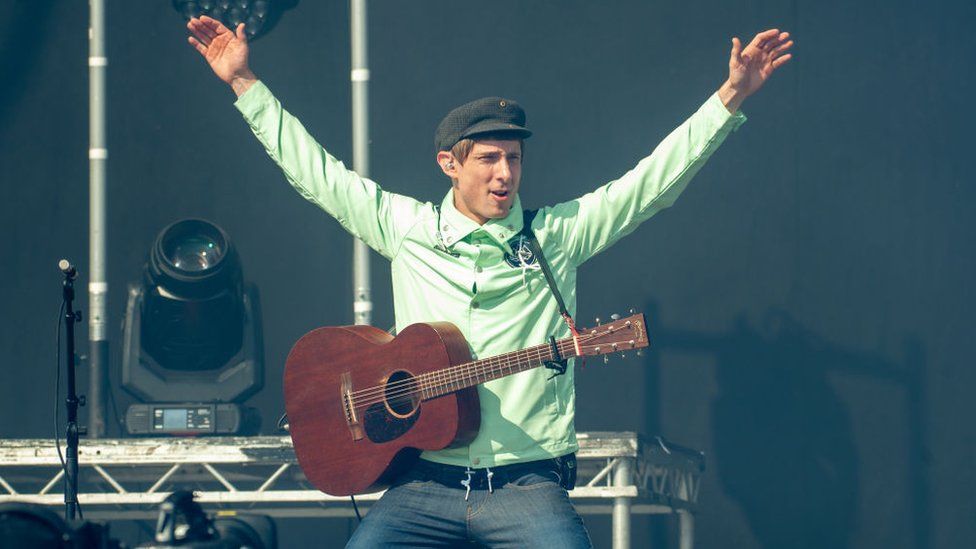 Gerry Cinnamon on stage at TRNSMT with an acoustic guitar, kneeling on the stage with his arms in the air