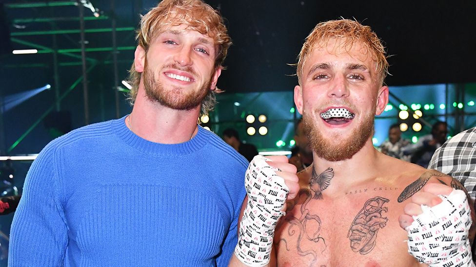 Logan Paul is on the left wearing a blue top and is smiling, while his brother Jake is on the right smiling at the camera with his fists point up in a fighter pose. The wrists and hands are taped up with a white bandage. The background has a few people, with some metal scaffolding pipes and spotlights.