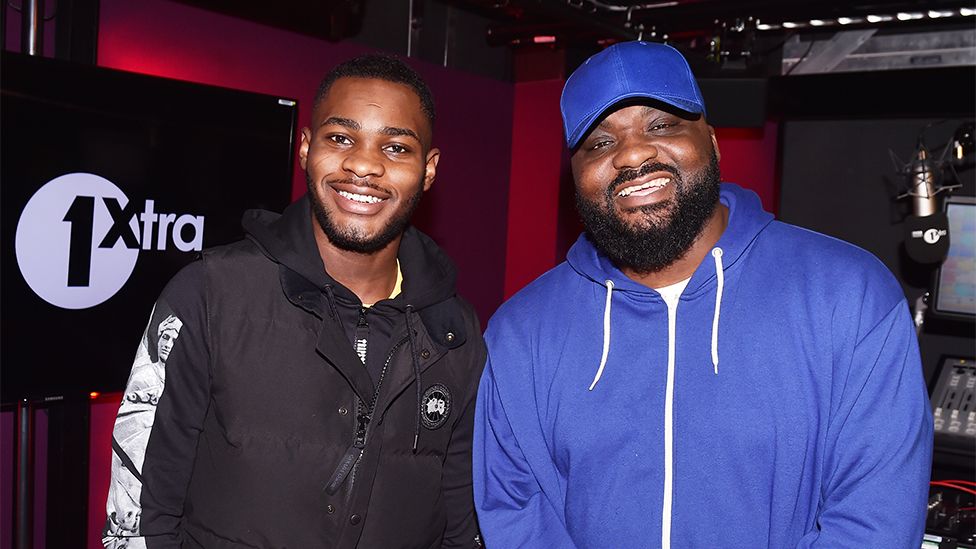 Dave and Ace. Dave on the left wearing a black tracksuit jacket smiling, with a 1Xtra sign on a black screen behind him. Ace on the right is smiling wearing a blue cap and hoody, with screens and buttons visible in the studio behind him.