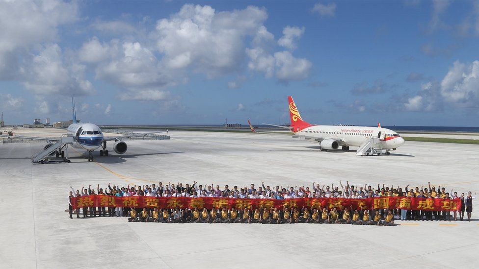 A large group of people pose for a group photo, carrying a large banner with yellow lettering on a red background, in front of two Chinese passenger jets on the airfield of Fiery Cross Reef, known as Yongshu Reef in Chinese. Taken 6 January 2016.