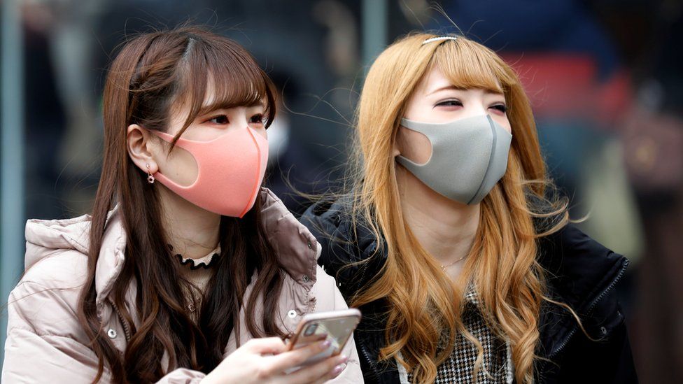 Two young women wear face masks in the street in Tokyo, in tones of pink and light grey