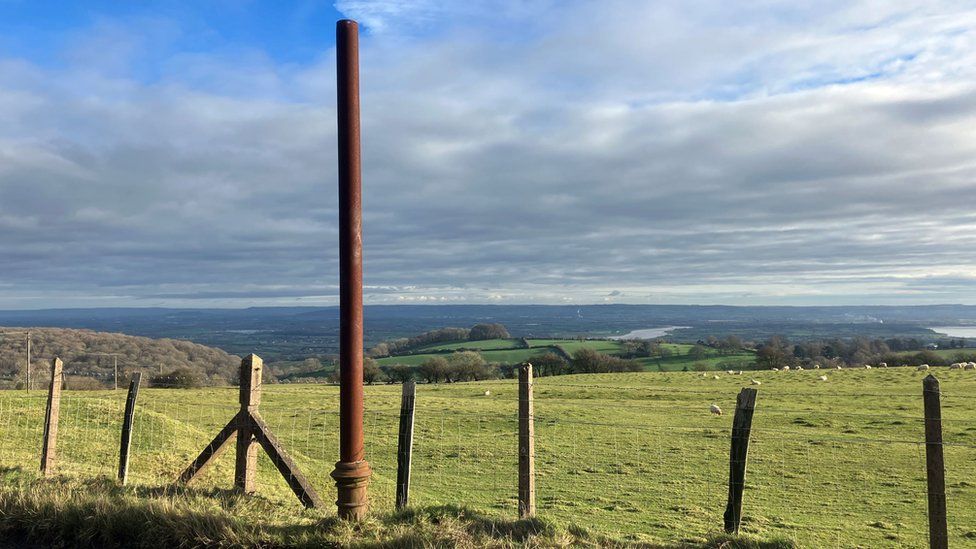 A large rusty pole on the side of a road next to a field of sheep