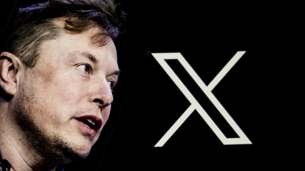 Elon Musk's photo is displayed on a phone screen in front of a computer screen displaying the new logo of 'Twitter'.