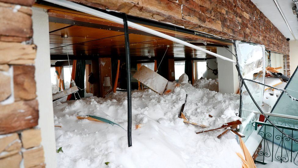 Snow is seen covering the inside room of a hotel, piled high enough to touch the ceiling lampshades, while the windows are shattered and broken