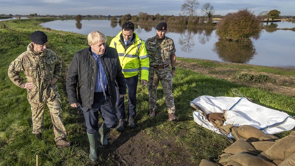 Boris Johnson walking along a flooded bank wearing wellies and surrounded by two soldiers
