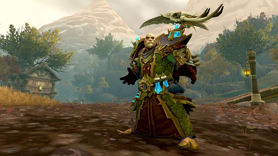 A World of Warcraft character with a bald head, bushy beard and black circles around his eyes stands on a muddy slope. He's wearing an ornate suit of armour with a large golden buckle at his waist. He has giant shoulder pads topped with blue jewels, and what looks like an eagle-like creature with its wings spread on his left shoulder. Two bright blue items - possibly potion bottles, hang from his belt. There's an old-world wooden cart or hut with ornate wooden trim in the background. There's a wooden post with a lantern hanging off it on the opposite side. The rest of the landscape is .ted with trees and tall, snow-covered mountains.