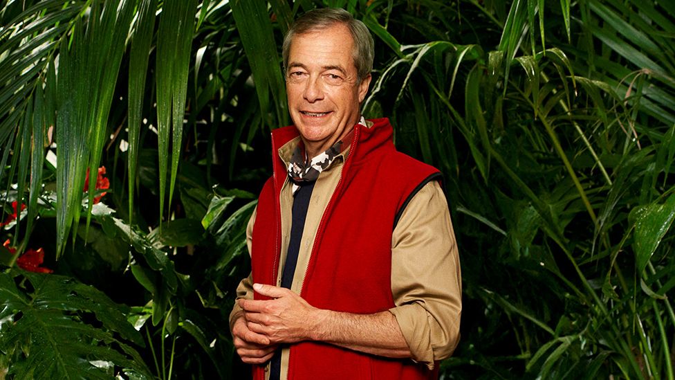 A man with short grey hair stands in front of a jungle background of dense foliage, made up of long, green leaves that hang down around him. He's standing with his hands clasped together, and wears a khaki (light brown) shirt with a red waistcoat/body warmer over the top. He's got a neckerchief on with a black, white and burgundy camouflage pattern.