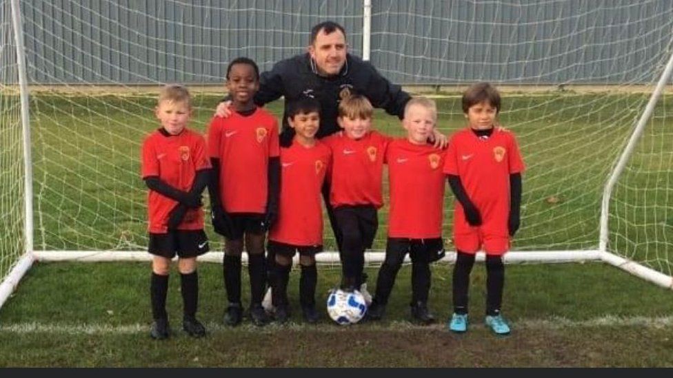 Matthew Evans standing in a football next with his arms around six young boys in their matching red and black football kit