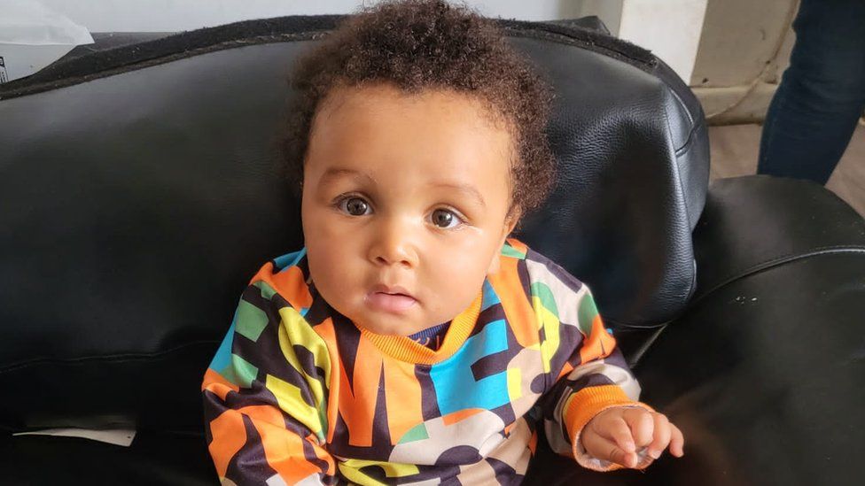 Baby Adonis looking at the camera with his hand outstretched, in a brightly coloured top.