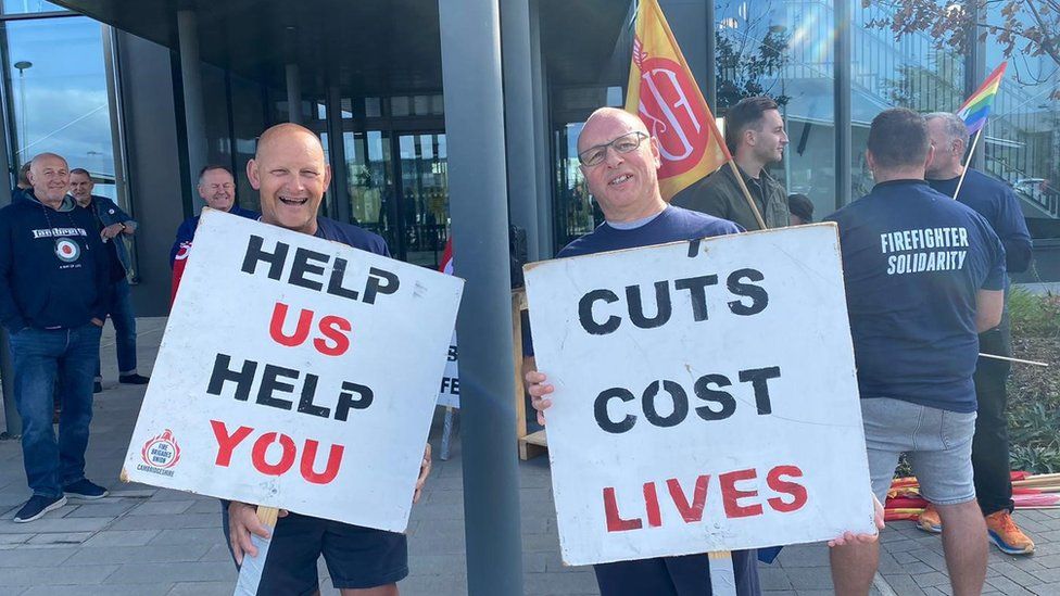 Two FBU members holding placards saying "help us help you" and "cuts cost lives"