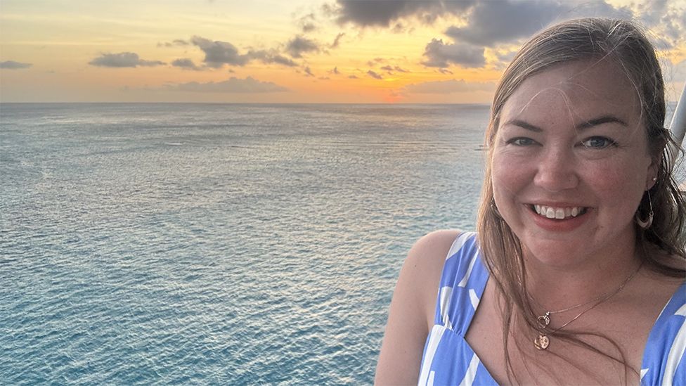 Angela Linderman smiling at the camera in a selfie, she is wearing a blue dress and her hair is down. She is in front of the sea