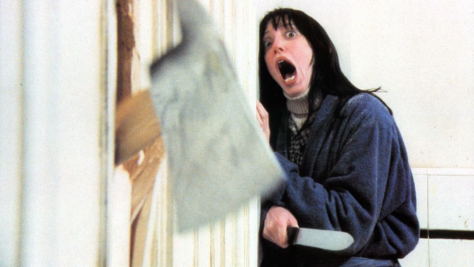 Shelley Duvall screaming as an axe comes through a door in a shot from The Shining