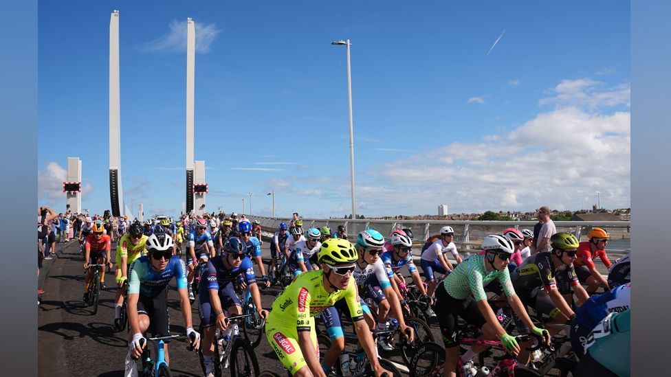 A large number of cyclists wearing colourful helmets and outfits race across gull wing bridge which has a huge white U-shaped monument at one end