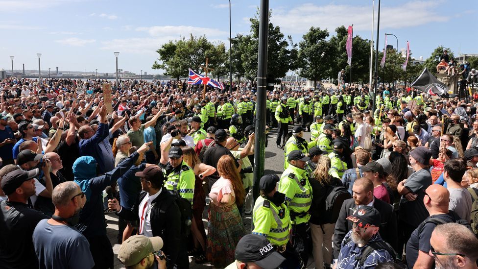 Two large groups of protesters face each other with two lines of police in hi-vis jackets standing between them