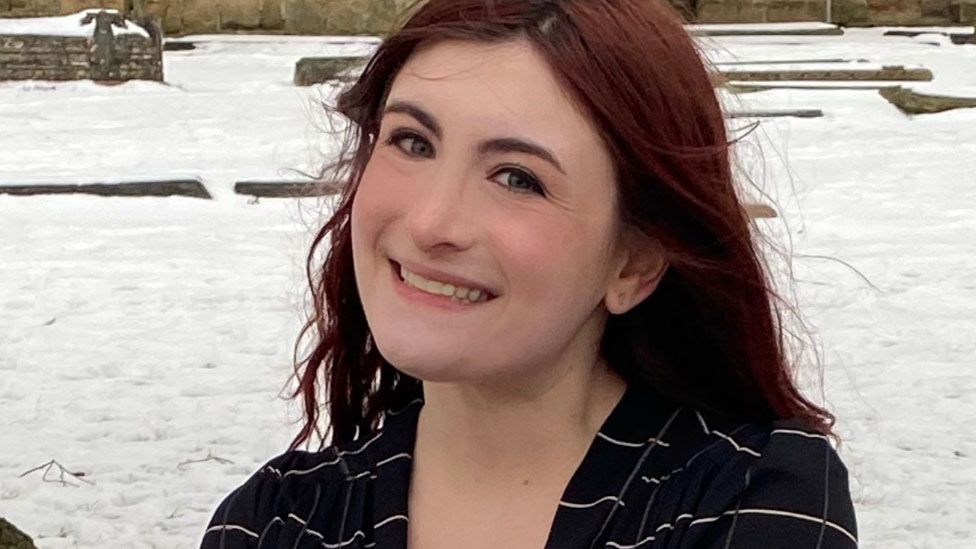 A woman with reddish brown hair wearing a black top with white cross lines looking towards the camera against a snowy backdrop