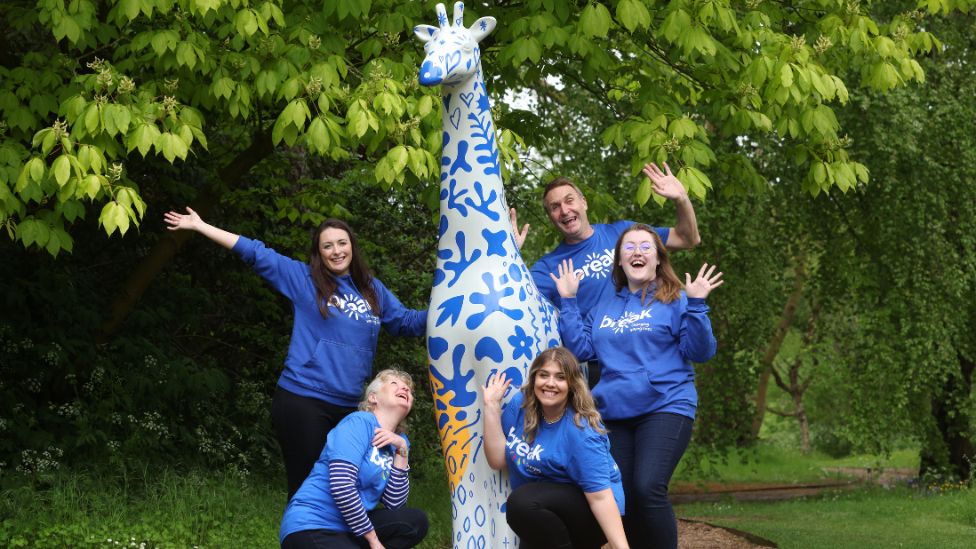 A giraffe sculpture decorated in blue and white and surrounded by five people wearing charity Break's blue fleeces