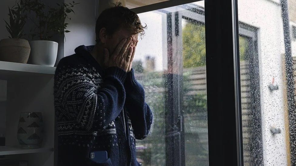 Man holding his face in his hands in sorrow, next to a rainy french window