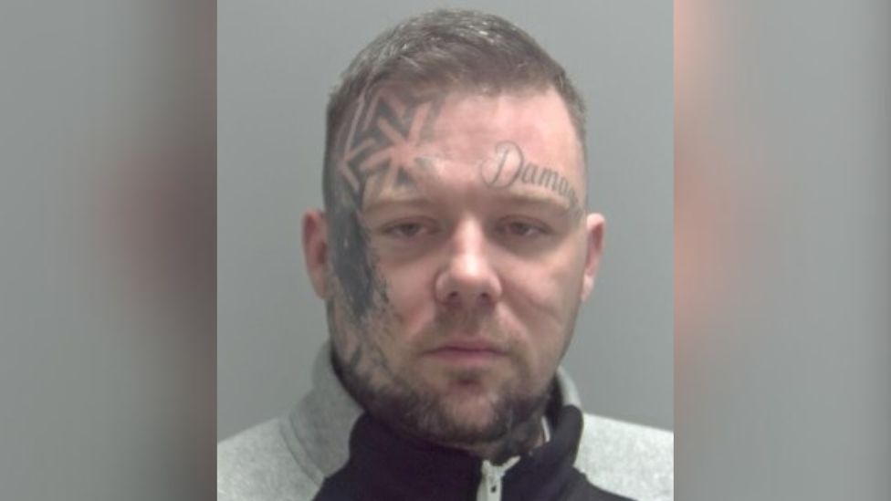 Custody mugshot of Daryl Taylor, he is wearing a grey and black sweatshirt and has large facial tattoos on the right side of his face and above his left eyebrow. 