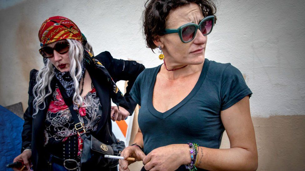 Two woman who described using drugs, including fentanyl. One wears sunglasses and a blue t-shirt and is holding a cigarette. The other wears sunglasses, a long black coat, and a red bandana.