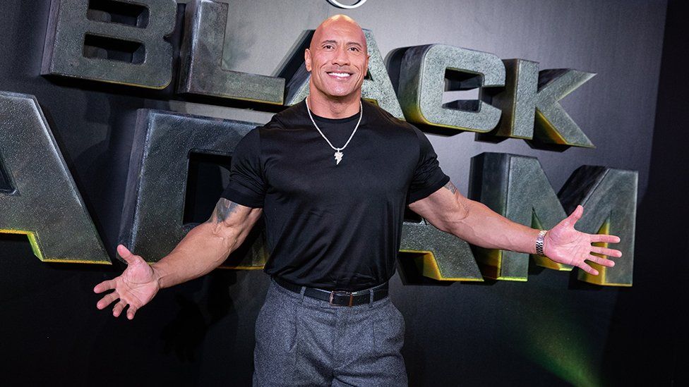 Dwayne The Rock Johnson pictured at a film premier. Johnson, a mixed race man in his 50s, has a shaved head and wears a black T-shirt which is tight over his muscly arms. He wears grey trousers and a diamond necklace. He has both arms outstretched as he smiles at the camera.