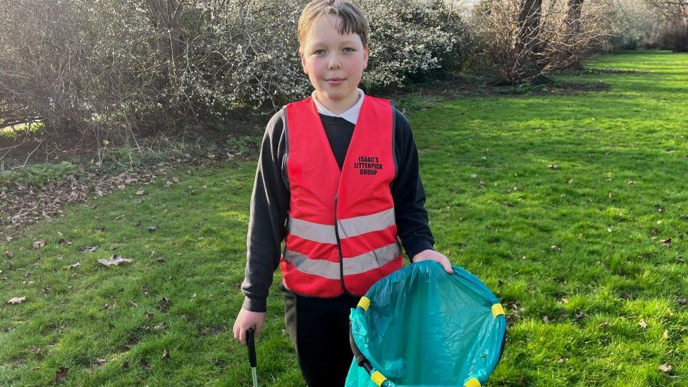 Boy wearing hi-viz vest with "Isaac's Litterpick Group" written on it, He is standing in a park with a litter picker.