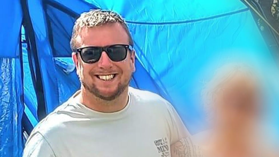 John Godwin smiles into the camera wearing sunglasses and a grey T-shirt