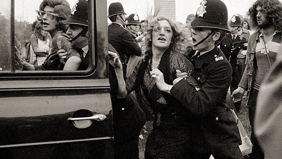 a drag queen is bundled into a 1970s police car