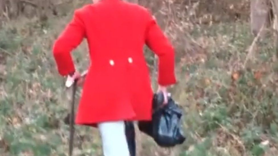 A man dressed in a red hunting jacket carrying a black bin bag