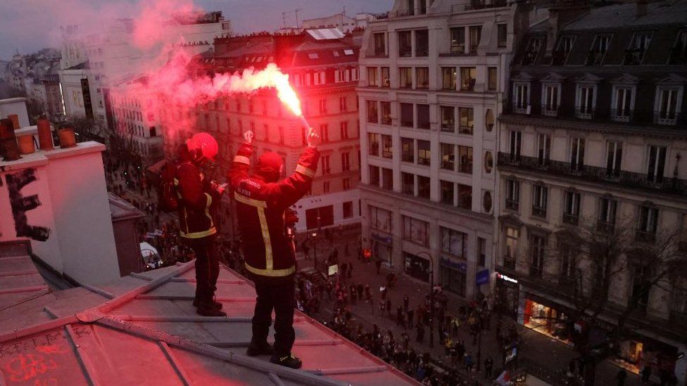 French firefighters on strike hold red flares as they stand on the top of a Parisian building during protests