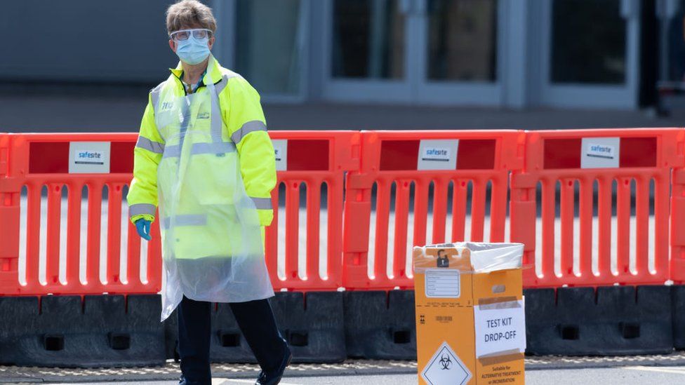 CARDIFF, WALES - APRIL 07: A worker wearing a surgical mask and apron stands near a test kit drop-off area at a drive-through testing centre at the Cardiff City stadium on April 7, 2020, in Cardiff, Wales. The testing centre opened today for frontline staff and people in the Gwent area. There have been over 50,000 reported cases of the COVID-19 coronavirus in the United Kingdom and over 5,000 deaths. The country is in its third week of lockdown measures aimed at slowing the spread of the virus.