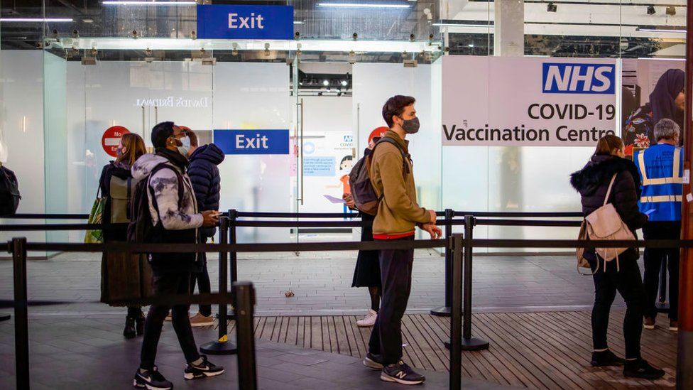 People are seen in a queue for their booster dose while standing at social distance outside the NHS vaccination centre at Westfield Stratford