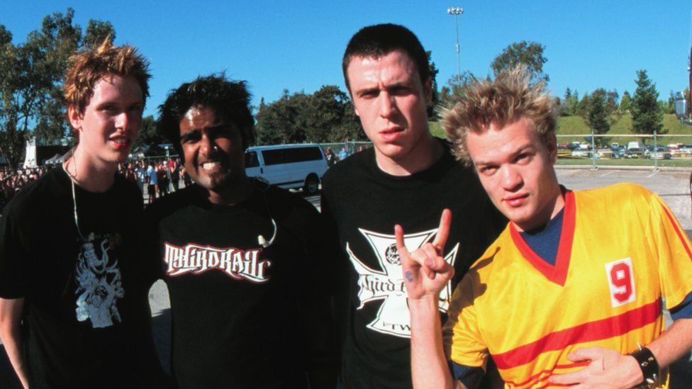 Sum 41 pictured in 2001. The band at the time was made up of (L-R) Jason McCaslin, Dave Baksh, Steve Jocz and Deryck Whibley. The band are in their early 20s and are photographed outside in a car park on a blue-sky day. Jason is a white man with red hair wearing a black T-shirt. Dave is an Asian Canadian man with grown out black hair and a nose piercing and a black T-shirt. Steve is a white man with cropped dark hair, also wearing a black T-shirt. Deryck is a white man with spiky blonde hair, wearing a yellow T-shirt with a red stripe.