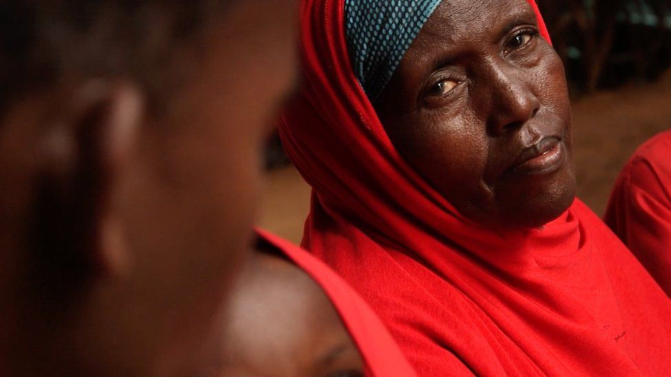 A woman dressed in red looks at the camera while she rests. A blurred child's face is in the foreground.