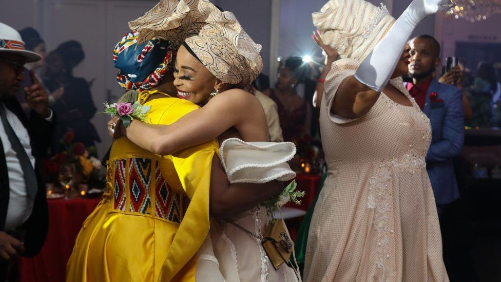 Two women at the "Bridgerton Affair" party hugging. They are both dressed up in party clothes and wearing headwraps. One of the women looks very happy and is smiling widely.