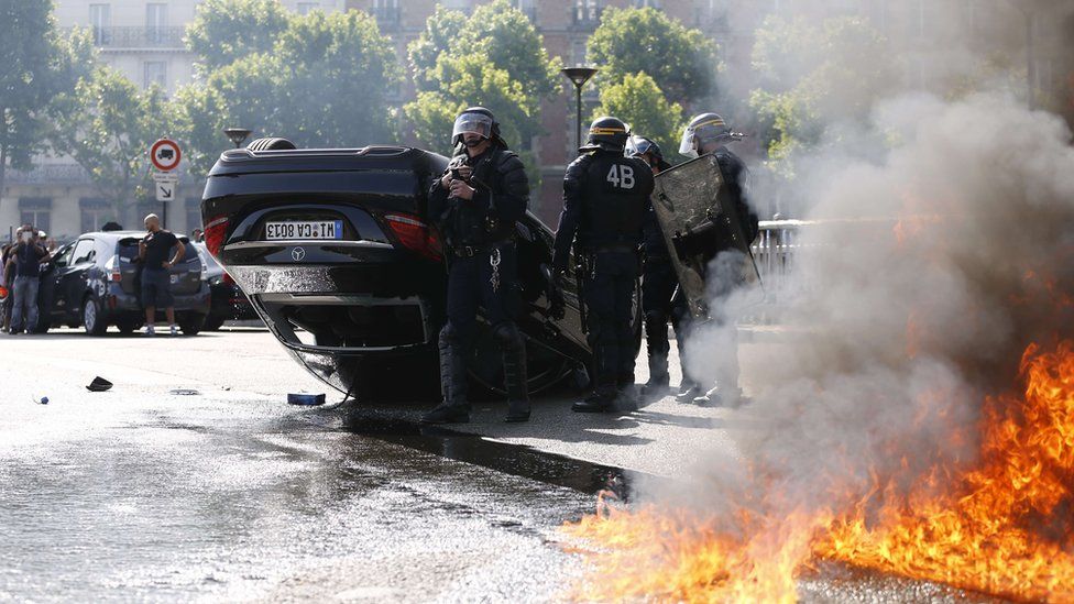Smoke rises from a fire burning next to French CRS riot police standing near an overturned car as taxi drivers block Porte Maillot in Paris on June 25, 2015.